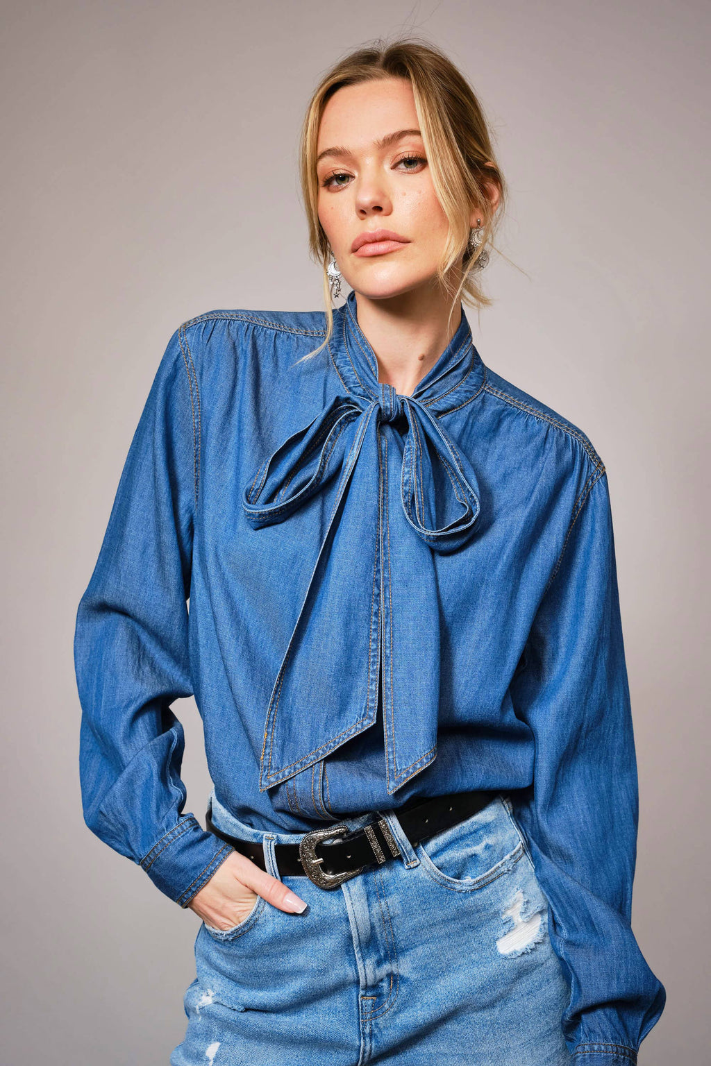 Woman wearing a blue denim shirt with a tie detail and matching jeans on a gray background