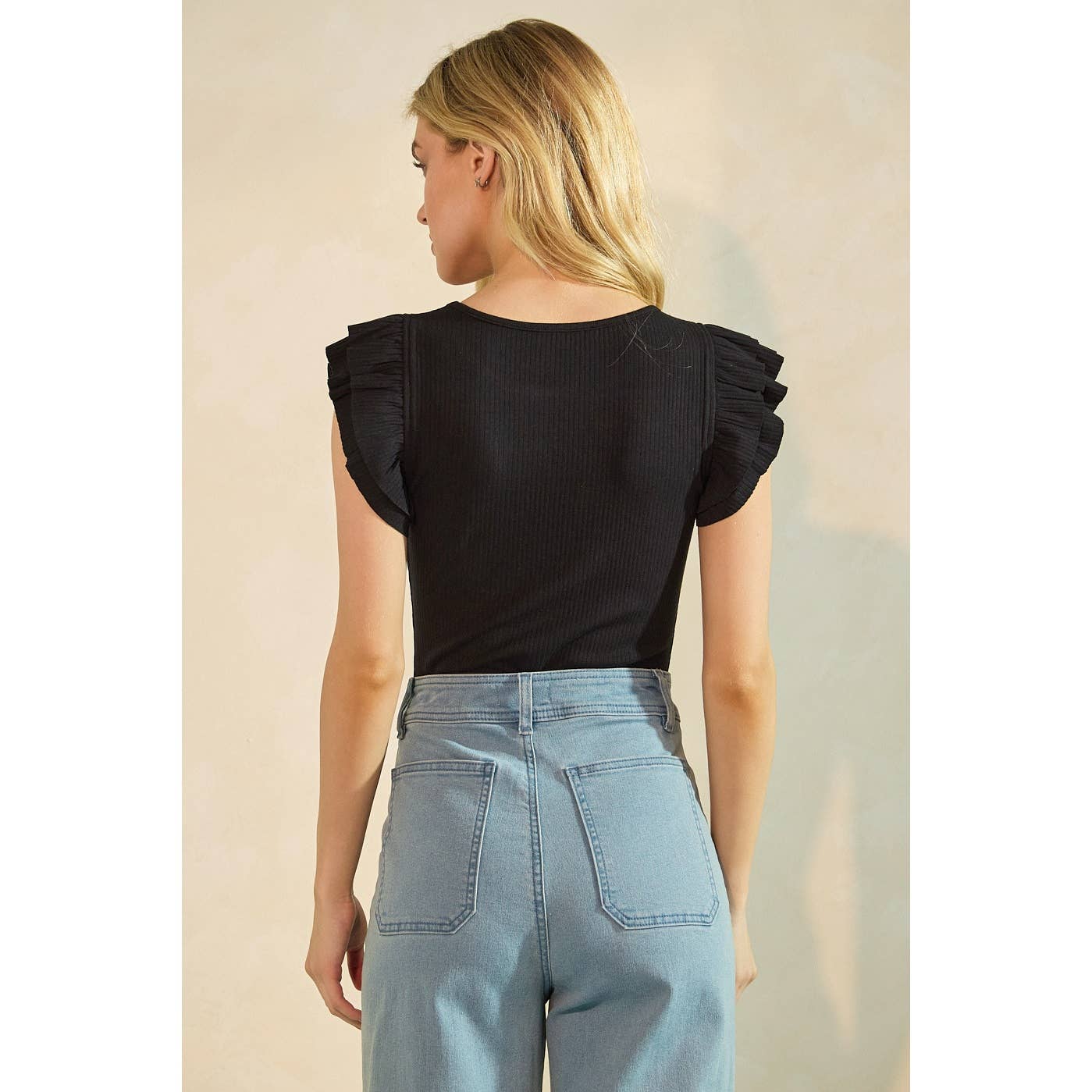 Woman wearing a black top with ruffled sleeves and light blue jeans against a neutral background