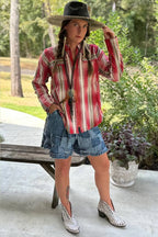 A woman modeling a red and white vertically striped western shirt, denim shorts, a black cowboy hat, and white western boots, standing outdoors on a concrete patio next to a wooden bench.