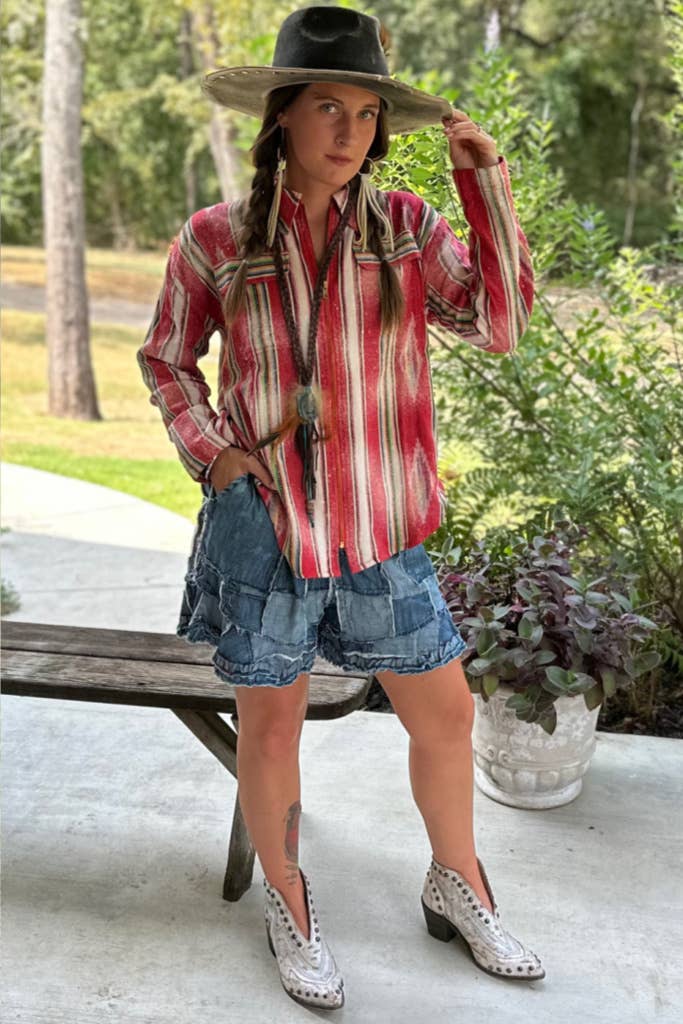 A woman modeling a red and white vertically striped western shirt, denim shorts, a black cowboy hat, and white western boots, standing outdoors on a concrete patio next to a wooden bench.