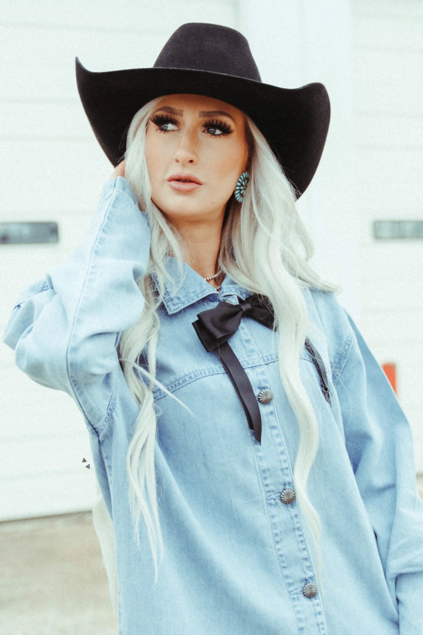 A woman wearing a black cowboy hat and a blue denim button-up shirt with the sleeves rolled up, styled with a black ribbon tied at the collar.