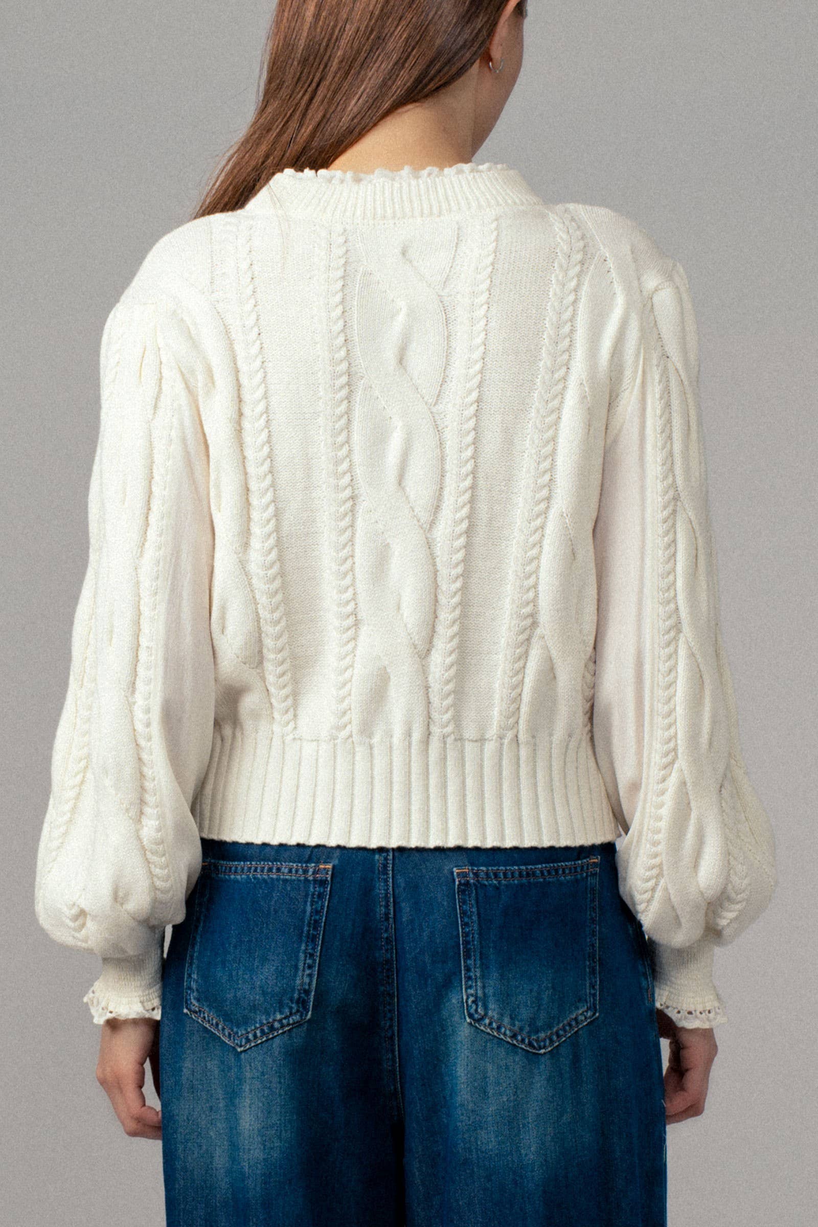 Person wearing a white cable knit sweater and blue jeans on a gray background