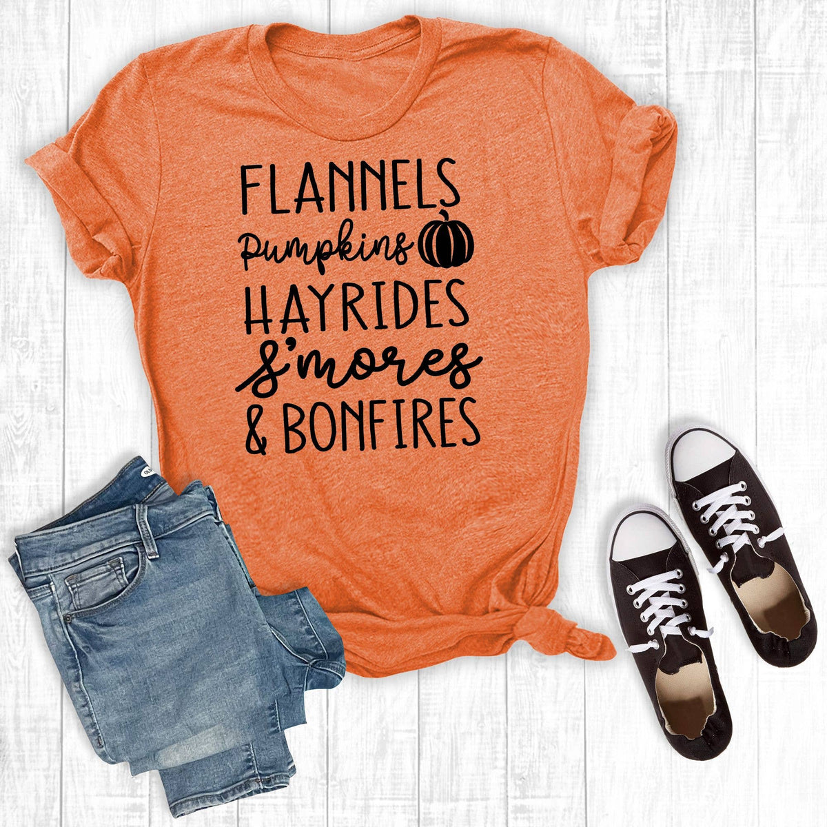 An orange heather graphic T-shirt laid flat on a white wood background, styled with folded blue jeans and black low-top sneakers. The shirt has black text reading 'FLANNELS Pumpkins HAYRIDES S'mores & BONFIRES' with a small pumpkin icon.