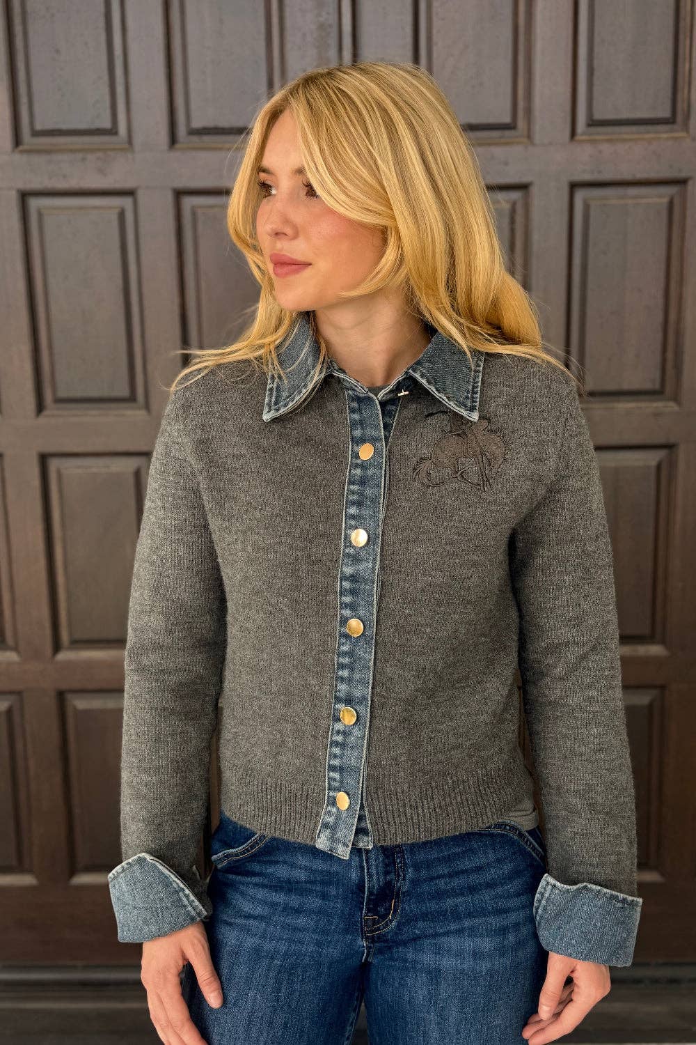 A woman wearing a gray knit cardigan with denim collar, cuffs, and front placket, paired with blue jeans, standing in front of a dark brown paneled door.