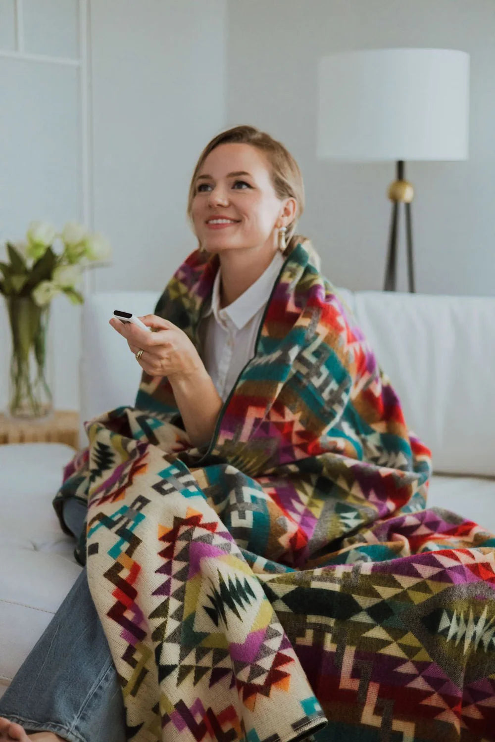 Woman sitting on a couch wrapped in a colorful patterned blanket, holding a phone.